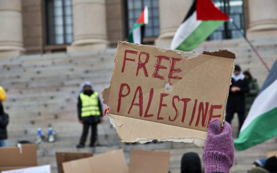 Photograph of a hand holding a cardboard sign above a protest, with handwritten block letters: Free Palestine. A palestinian flag waves in the background in front of a stone large building with pillars.