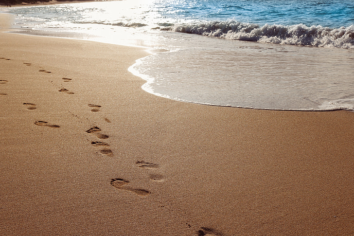 Footsteps on wet sand next to receding waves on a beach.